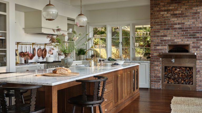 Cette ferme des années 1920 a été rénovée une fois de trop – jusqu’à ce qu’une rénovation réfléchie lui redonne vie A spacious, sunlit kitchen featuring a large oak island with a marble countertop, black spindle stools, and vintage-style glass pendant lights