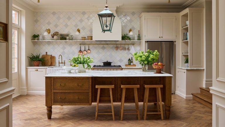 Vous pensez que les cuisines rose pâle seront disponibles en 2026 ? Cet espace plein de caractère prouve que ce neutre chaleureux est en fait la base idéale pour les plus grandes tendances de cette année A wide shot of an elegant kitchen with herringbone wood floors, featuring a white fluted farmhouse sink, brass hardware, and a marble-topped oak island. Natural light streams in from a large window on the left, illuminating the soft cream and grey diamond tiles