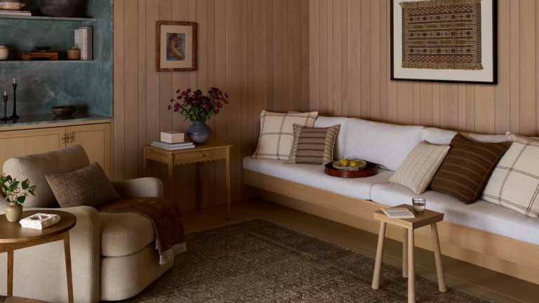 Amber Lewis est « obsédée par les détails » : ce sont les 5 éléments de décoration qu’elle trouve les plus chics et pourquoi vous devriez les utiliser dans votre maison a warm living room with a built-in couch with pillows on top, a brown swivel chair, a stool, and a side table with flowers on it