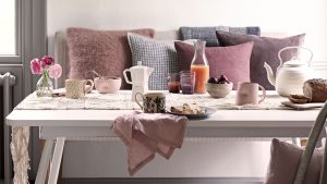 La nouvelle cafetière Moka à 10 $ d’ALDI est la mise à niveau du café au look design qui appartient à chaque comptoir de cuisine moderne A brightly lit breakfast scene features a white dining table covered with a cream macramé runner and an assortment of soft pink and patterned ceramics. On the table, there is a white teapot, a Moka pot, a carafe of juice, bowls of fruit and cereal, and a small glass vase with pink ranunculus. Behind the table, a bench is styled with several plush cushions in shades of dusty rose, mauve, and grey-blue. The overall aesthetic is airy and cozy, leaning into a soft, romantic color palette.