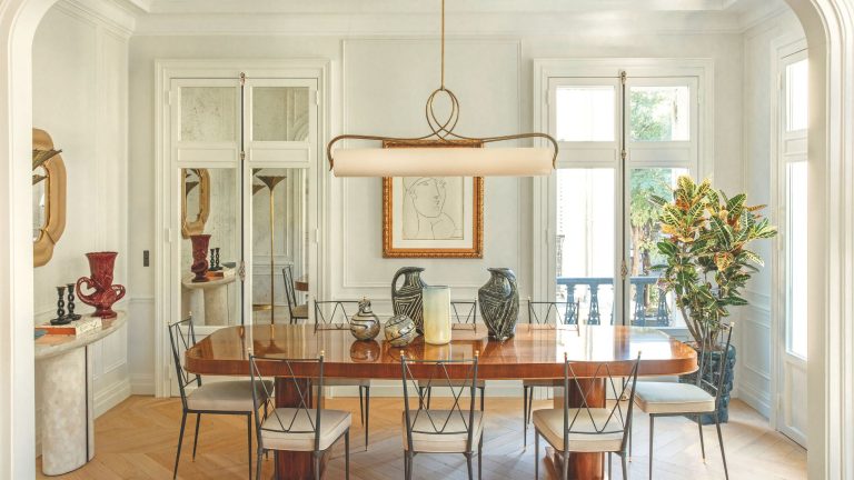 « Dès que vous remettez les pieds dans l’avenue Frochot, vous entendez le chant des oiseaux » – Explorez un appartement élégant au milieu de Paris, une évasion calme de l’agitation de la ville An elegant, sunlit dining room featuring a polished wood table, slender black metal chairs, and a unique horizontal pendant light. The room is decorated with ceramic vases and a large framed sketch on a paneled wall.
