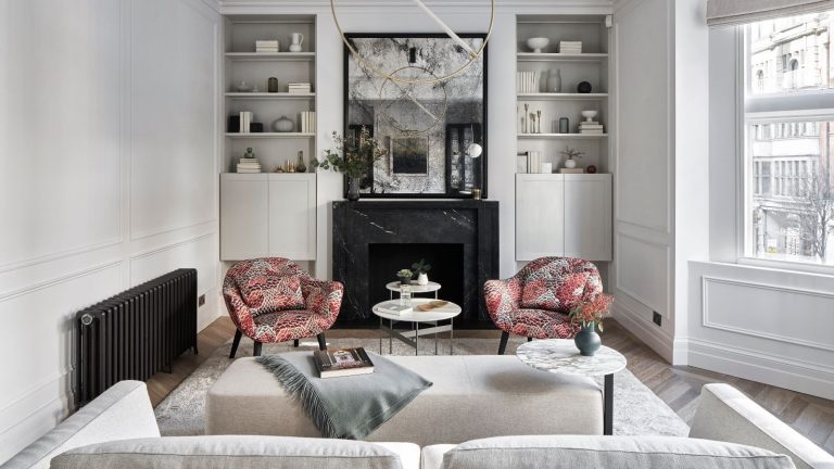 Réchauffez les pièces plus rapidement et gaspillez moins d’énergie grâce aux housses de plinthe chauffante à 3 $, révèlent les professionnels du CVC A white living room with white rug, white couch, white footrest, pink and red patterned chairs, a black radiator, and white shelving around a black marble fireplace with a black mirror above.