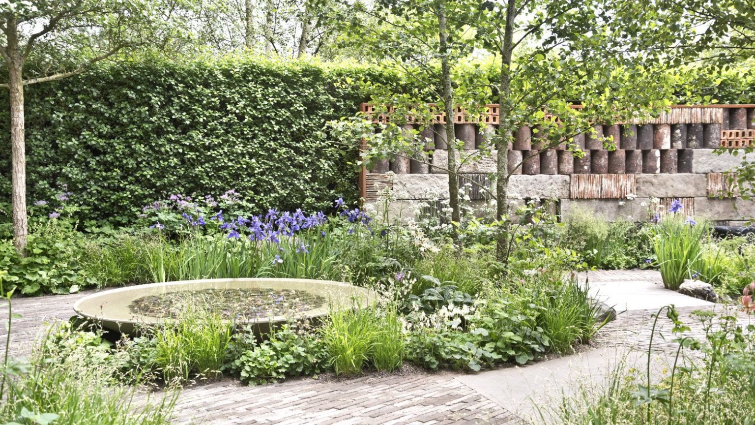 Ces éoliennes sont garanties pour apporter un peu de fantaisie à votre jardin – et éloigner les parasites de vos cultures. A show garden complete with a brick patio, circular water feature, borders, and a large hedge at the back