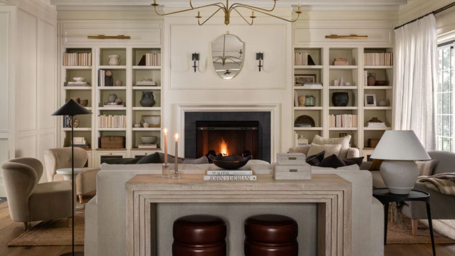 La symétrie est-elle démodée ? Les designers révèlent la formule d’aménagement du salon qui brise les règles et qui va redéfinir la façon dont nous décorons en 2026 neutral living room designed by mcgee and co with lit fire, symmetrical alcove bookshelves, a neutral floating couch with travertine table and little stools