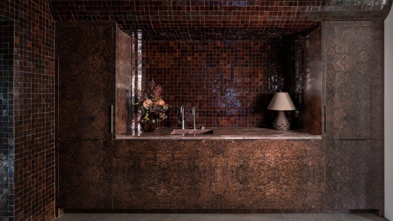 a bar area with burl wood cabinetry, dark brown zellige tile, a sink, and a vase of flowers on the countertop