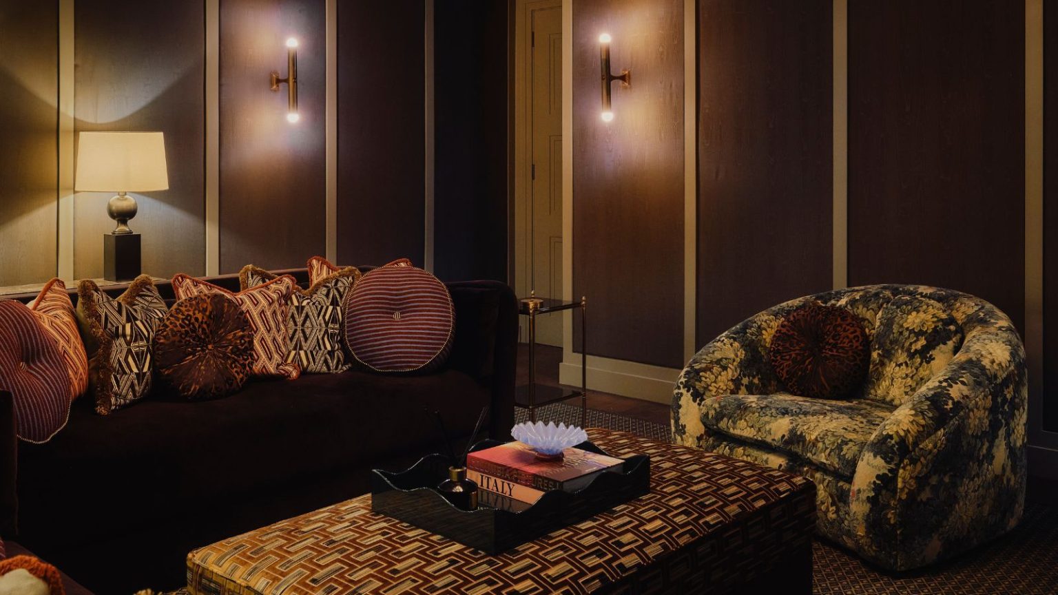 dark brown snug room with dark couch, patterned throw pillows, a tapestry print arm chair, and velvet upholstered ottoman