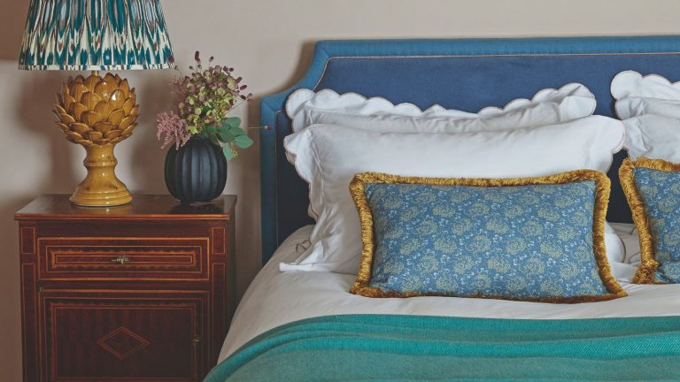 Ce « piège à poussière » domestique courant aggrave vos allergies – mais un accessoire d’aspirateur à 11 $ peut facilement le réparer Cream bedroom with blue headboard and white bedding with a wooden bedside table and yellow lamp
