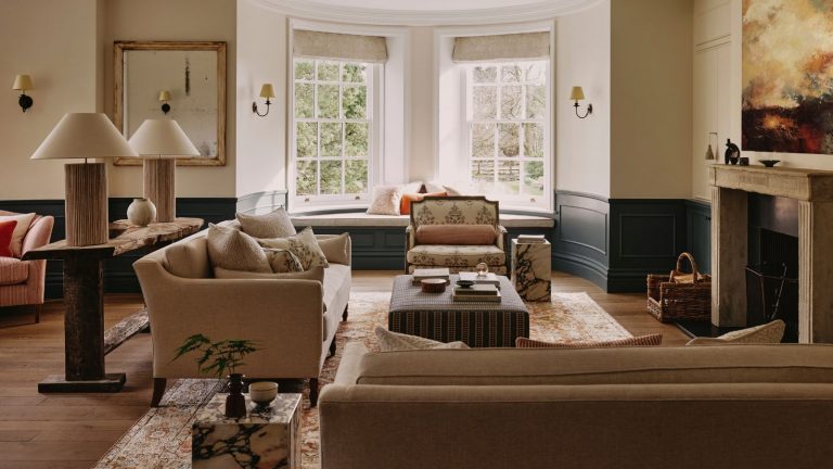 Parfois, ce que vous ne mettez pas dans votre salon compte autant que ce que vous faites – les designers expliquent le pouvoir de l’espace négatif neutral living room with original stone fireplace, two tone half panel in navy blue, neutral couches, a couch table with two large lamps and a striped ottoman
