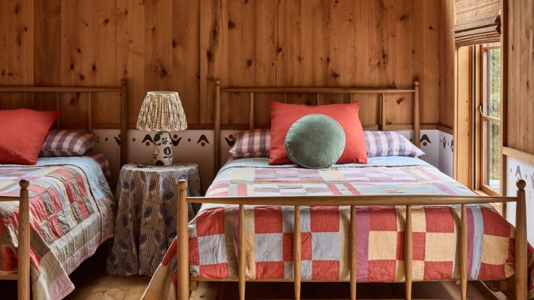 Better Vintage : « Un patchwork de lieux et de mémoire » – Pourquoi les courtepointes vintage restent des objets de collection, pratiques et profondément personnels Rustic twin bedroom with wood paneling, sloped ceiling, an A-frame window overlooking trees, and bright patchwork quilts