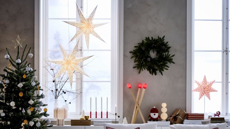 Je n’ai pas non plus pu obtenir les étoiles STRÅLA d’IKEA cette année, mais ces alternatives chics de style scandinave apportent le même éclat élégant et intemporel à ma maison IKEA paper stars hung at the window of a living room with gray walls, a wreath between the window and a christmas tree decorated with white and gold