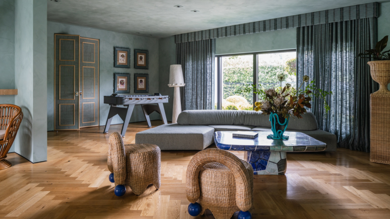 Adieu les rideaux ennuyeux, la tendance autrefois démodée des habillages de fenêtre luxueusement spectaculaires est de retour – et les concepteurs l’approuvent tout à fait. a large living room with light blue painted walls and ceiling, blue curtains blocking the large open windows, a blue couch, and wicker accent chairs