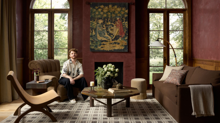 L’arbre de Noël « collecté » de Lone Fox est à couper le souffle – et je viens de trouver tout ce dont vous avez besoin pour recréer son look douillet et superposé a moody, medieval-inspired living room with a brown and gray checker printed rug, a chocolate brown sofa, two accent chairs, red painted walls, a vintage-inspired tapestry, and a man sitting on one of the chairs