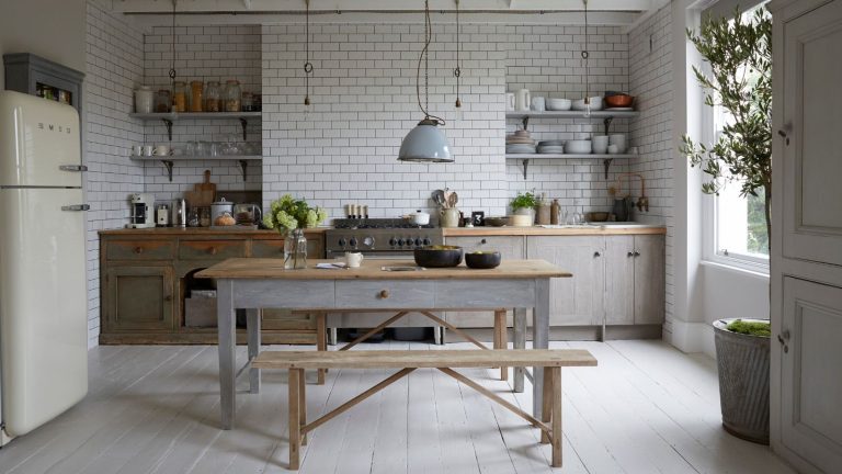 « Arrêtez de traiter votre comptoir de cuisine comme une table basse envahie » : voici comment les concepteurs équilibrent l’ajout de caractère sans ajouter d’encombrement a large kitchen with a freestanding table with a bench in the center, a wall of countertops and base cabinetry, shelfs on the tiled wall, and a hanging pendant light