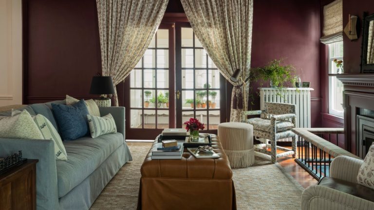 Oubliez la télévision – Cet aménagement de salon a été entièrement conçu pour la conversation avec une idée de canapé flottant que vous voudrez copier dark reddy purple color drenched living room dressed with a light blue skirted couch, skirted leather ottoman, floral armchairs and floral drapes