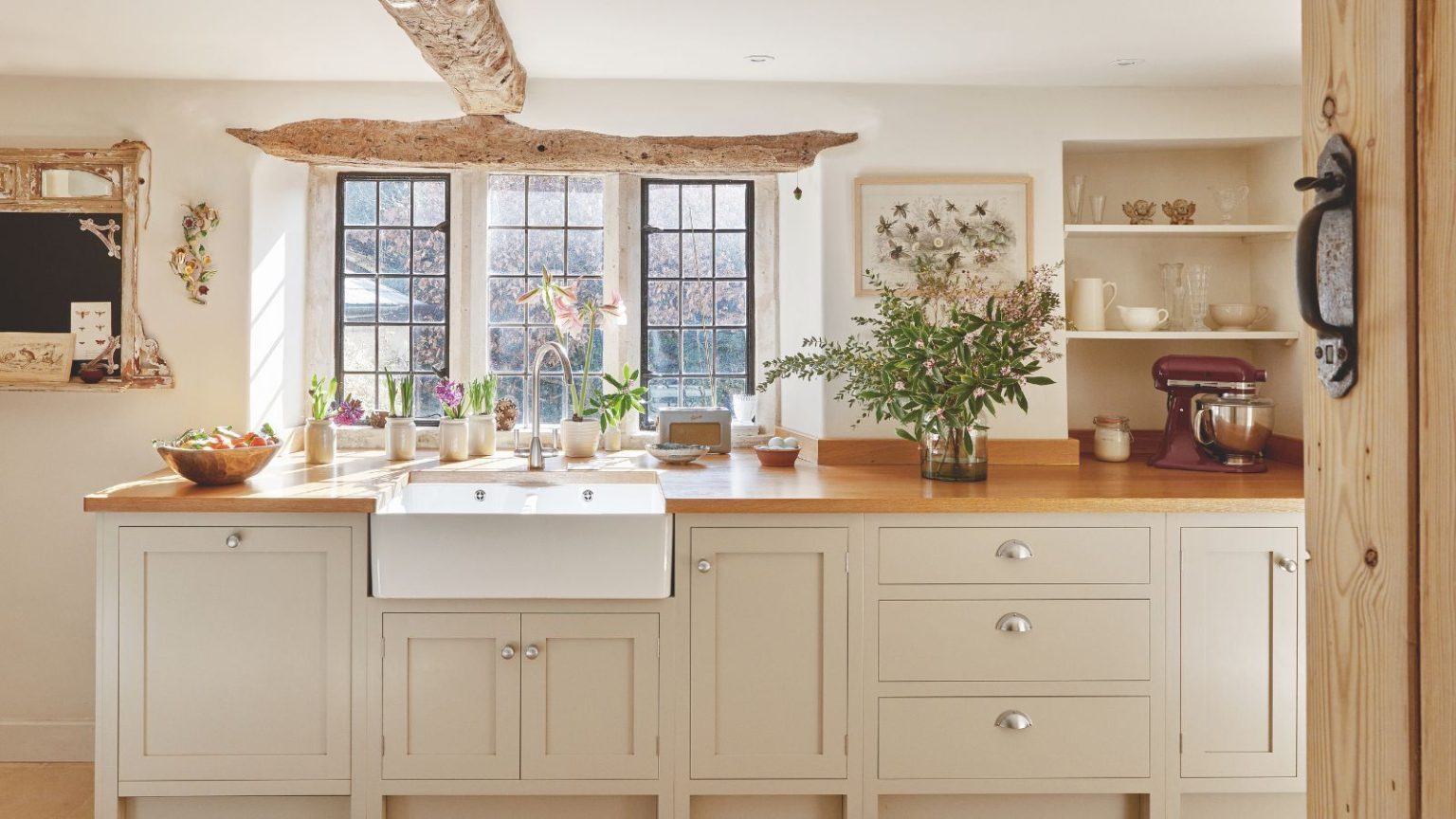 24 découvertes de rangement de cuisine Amazon qui résolvent instantanément les problèmes d’organisation de cuisine les plus courants A bright and open country-style cream kitchen with a white farmhouse sink set into a wooden counter below a bright window. Gray painted cabinetry with silver hardware is visible, as are original wooden beams on the ceiling and above the crittal window.