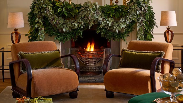 L’édition très britannique : chaleureuse, lumineuse et confortable – Comment le style au coin du feu transforme les pièces en retraites cocooning et prêtes pour l’hiver Cozy, warmly lit living room centered around a stone fireplace decorated for Christmas. The hearth contains a roaring fire and is framed by a garland. To the right, a comfortable club chair sits next to a wooden coffee table with a table lamp. The room features a neutral palette with natural materials, throws, and festive decorations including a houseplant and holiday accents.