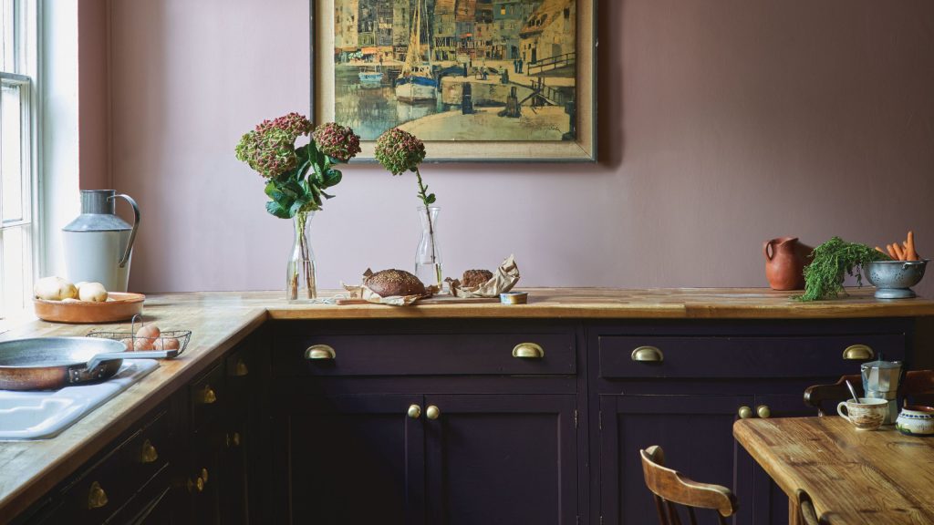 Farrow &amp; Ball kitchen with a pink wall and dark purple cabinetry with wooden worksurface and rustic wood dining table and chair