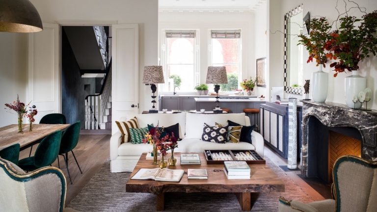 An eclectic living and dining space with a wooden dining table and dark green velvet chairs to the left, and a large wooden coffee table in the centre to the right, with a large rug and white couch with patterned, mismatched cushions. To the right is a black and white marble fireplace with two white vases on top. On the back wall are two large windows, and two matching lamps.
