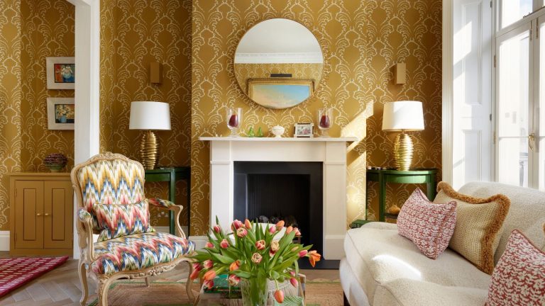 Déplacez-vous sur le jaune beurre : l’ocre est le neutre douillet que les designers adorent cet automne A traditional living room with ochre and white patterned wallpaper on the walls, a fireplace with a round mirror above, and a cream sofa with ochre and red cushions.
