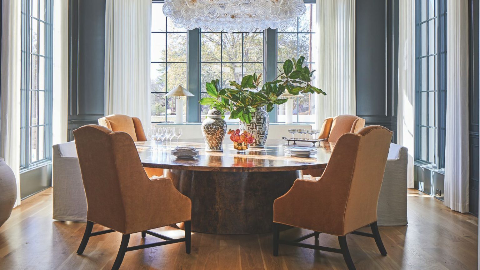 Le détail de conception par lequel cette décoratrice d’intérieur ne jure que a donné à son tout nouveau projet de maison une impression de caractère et de « vraiment sur mesure » Blue paneled dining room with large windows and long white drapes. A circular polished wooden dining table sits in the middle with four orange upholstered chairs around it