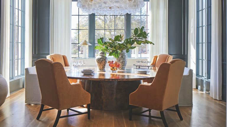 Le détail de conception par lequel cette décoratrice d’intérieur ne jure que a donné à son tout nouveau projet de maison une impression de caractère et de « vraiment sur mesure » Blue paneled dining room with large windows and long white drapes. A circular polished wooden dining table sits in the middle with four orange upholstered chairs around it
