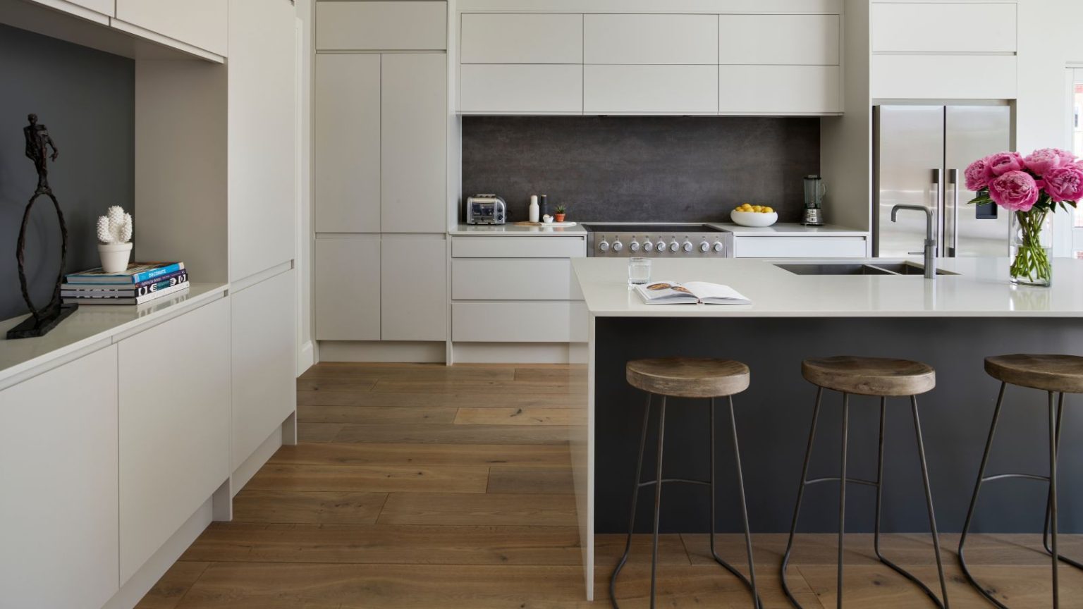 Vous envisagez de relooker votre cuisine ? N’oubliez pas cet élément essentiel du stockage, préviennent les professionnels de l’organisation A modern white kitchen with white cabinets, wooden flooring, a dark grey backsplash, and a central white kitchen island with a dark grey space underneath the counter. At the counter are three matching metal and wood barstools, and on top of the counter is an open recipe book, glass of water, silver faucet with recessed sink, and a glass vase of large pink flowers.