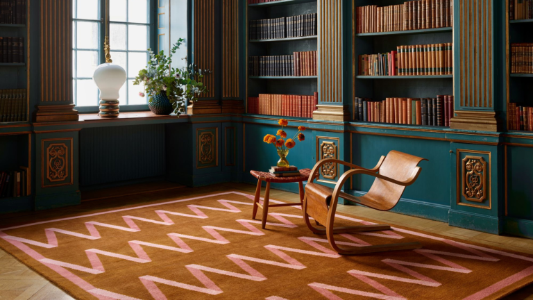 J’ai enfin identifié le tapis qui domine mon flux Instagram – Découvrez le tapis à ruban pliant et découvrez pourquoi les designers en sont obsédés a study room with a large rug with pink zig zag stripes and a brown background, teal painted bookshelves holding many books, and a wooden chair and an accent table in the middle of the room