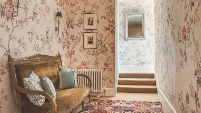A pink floral wallpapered hallway with a vintage upholstered bench alone one wall.