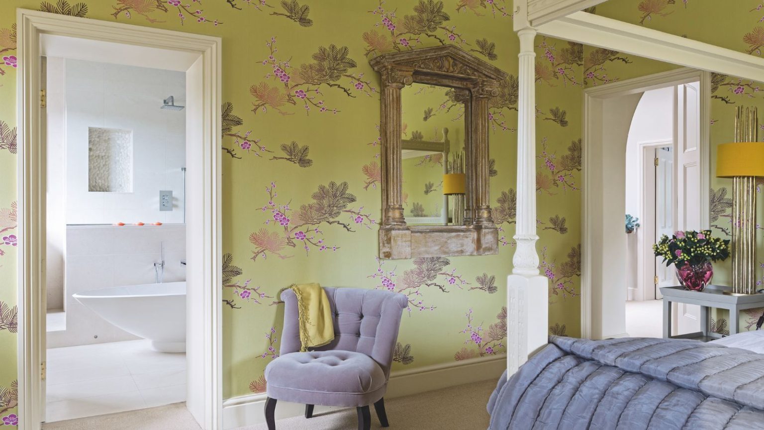 L’article de rangement secrètement toxique dans la salle de bain que les toxicologues disent que vous devriez remplacer cette saison A green bedroom with statement wallpaper and a white four-poster bed with a grey blanket. To the left is a doorway open to a white en-suite bathroom, and a grey upholstered chair.