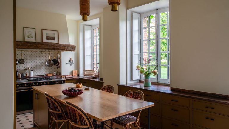 A country kitchen with deep red lower cabinets, and a large wooden dining table in the centre.