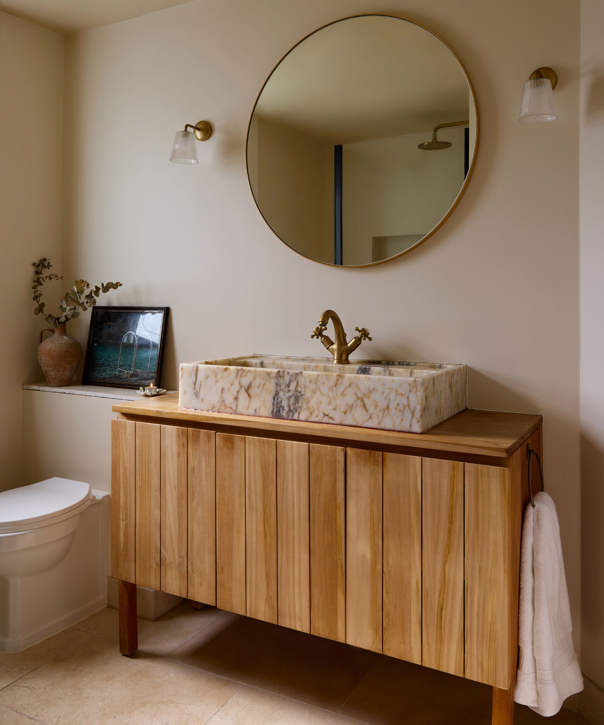 salle de bains moderne et confortable peinte en Farrow &amp; Ball's Dimity avec une vanité en bois chaleureux et un lavabo en marbre veiné rose, un miroir rond et des appliques murales en laiton