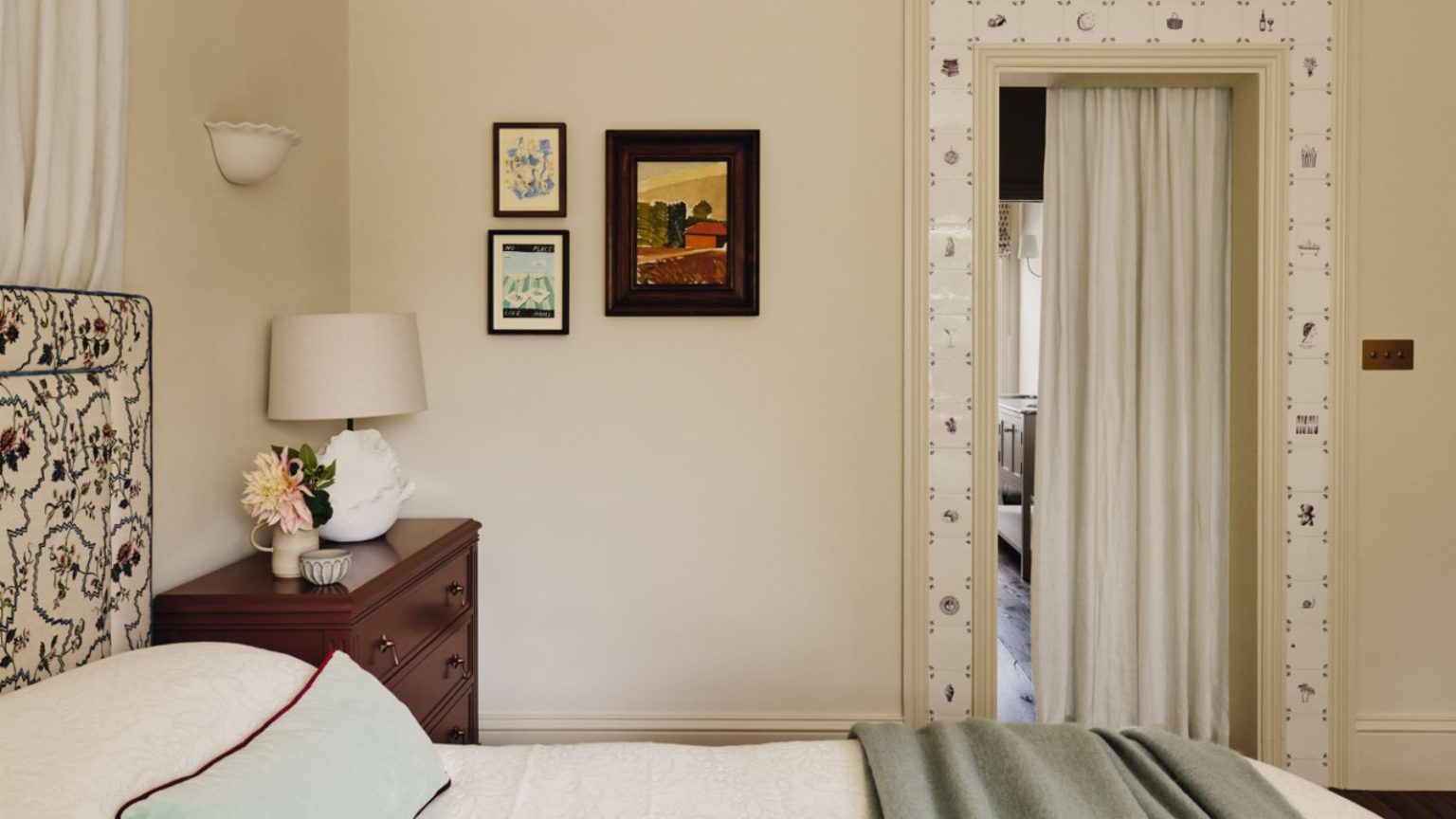 J’ai utilisé Dimity, le best-seller de Farrow & Ball, dans quatre pièces de ma maison. Voici pourquoi je conviens qu’il s’agit de la meilleure teinte neutre et chaude pour créer des espaces doux et apaisants. neutral bedroom painted in Farrow & Ball's Dimity with delft tile door trim, small vintage artwork, a floral upholstered headboard and a burgundy nightstand