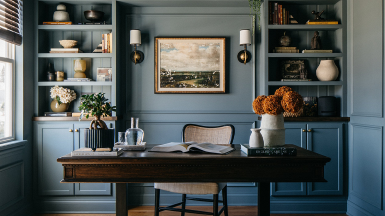 a home office drenched in a soft blue/gray color with built in shelves covered with decor, a freestanding desk with decor on top, a chair behind it, and a vintage-inspired rug on the floor