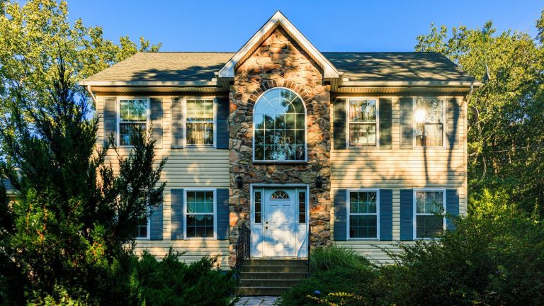 Les conditions météorologiques extrêmes ont révélé ces 3 points faibles coûteux pour votre maison en 2025 – Renforcez le vôtre maintenant avant qu’il ne soit trop tard Two-story suburban house with distinctive stone facade and arched window. Beige siding, blue shutters, and green roof harmonize with natural surroundings. Lush greenery frames property, mature trees and shrubs add privacy. Stone steps lead to white front door, creating inviting entrance.