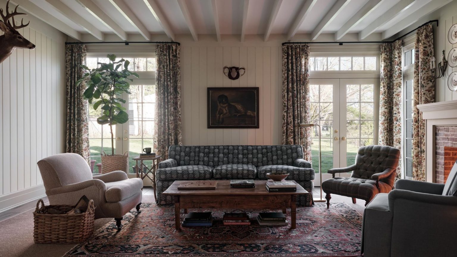 Vous n’avez pas besoin de vivre à la montagne pour avoir une « maison de montagne » – Voici comment recréer cette tendance nostalgique n’importe où a cozy living room with rustic influence. A large, patterned sofa sits against a wall with two sets of french doors, accent chairs are on either side of the couch, a coffee table sits in the middle of the room, and a deer head hangs on the wall