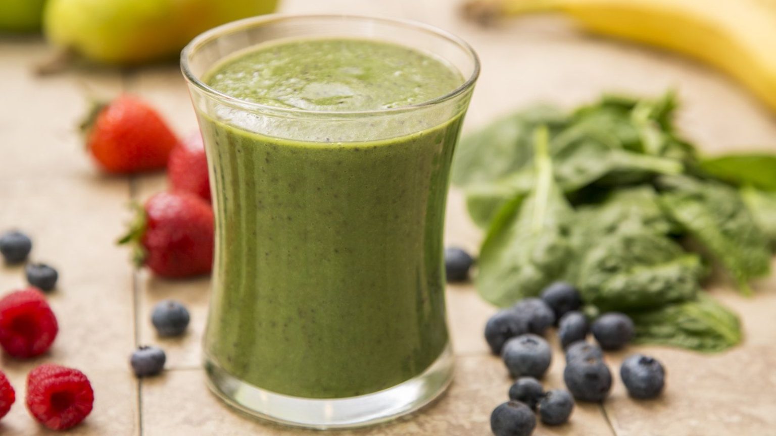 Commencez ces 7 cultures faciles dès maintenant pour des smoothies cultivés sur place d’ici l’été A green smoothie in a glass surrounded by spinach, blueberries, and strawberries on a table