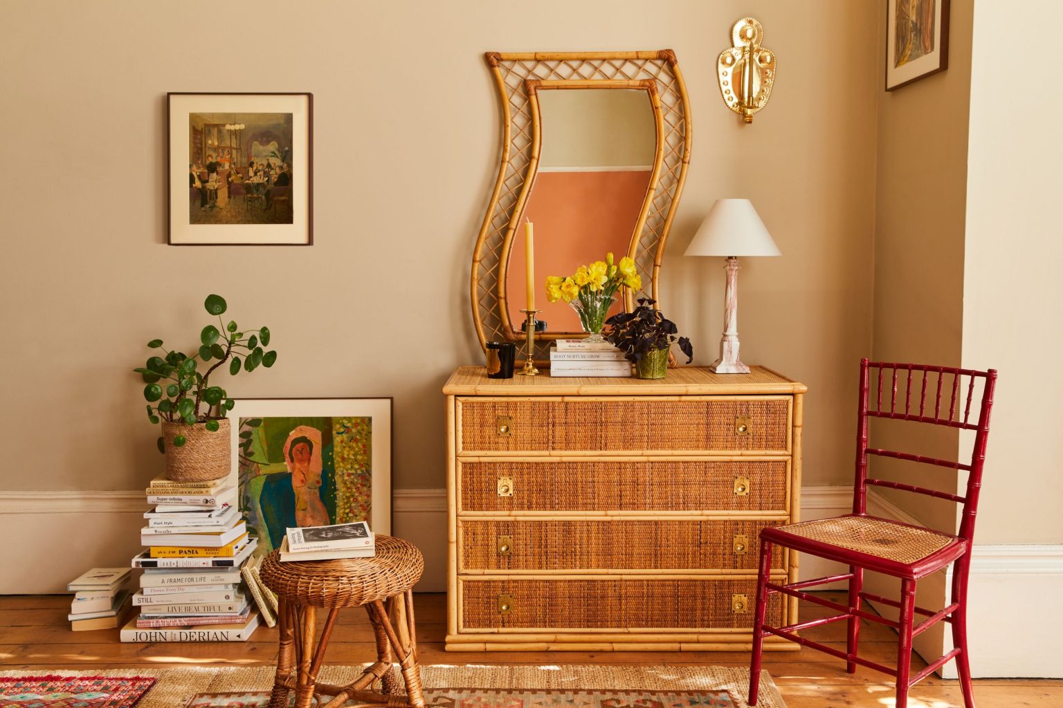 « C’est une petite décision qui donne discrètement le ton à une pièce » – Pourquoi l’éclairage vintage perdure-t-il dans les maisons modernes A textured bamboo dresser topped with a unique, wavy-framed rattan mirror, yellow daffodils in a glass vase, and a pink marble lamp against a beige wall.