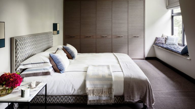 J’ai presque gâché l’heure d’un film – jusqu’à ce que cette astuce de barre de son transforme nos soirées : des dialogues nets, un volume doux et un calme total sans réveiller la maison A neutral bedroom space with large brown closets, a window seat with cushions, and a large double bed with patterned headboard, white bedding, and cushions, and a marble bedside table beside it.