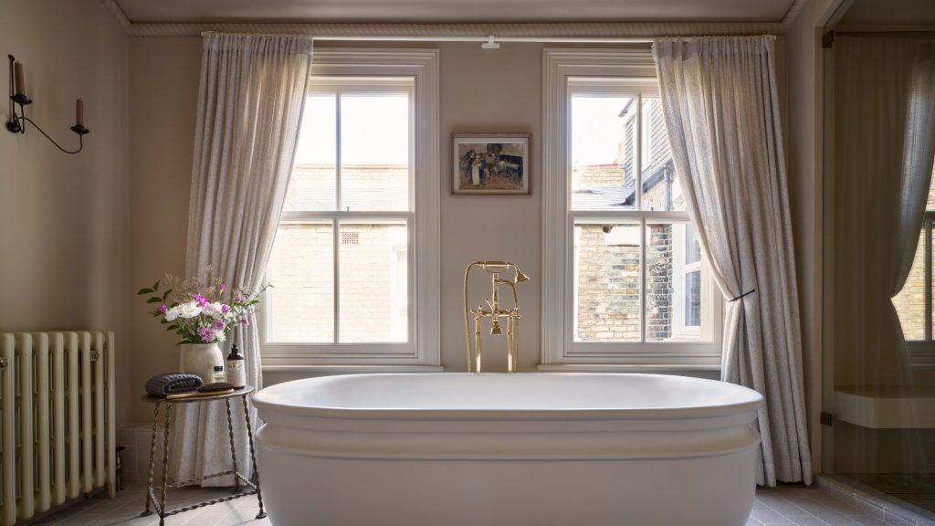 A warm neutral bathroom with a freestanding bath and floor length curtains over the windows