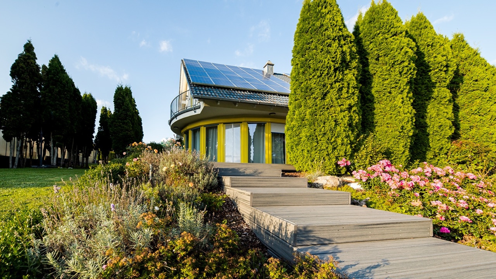 Photo lumineuse d'un escalier en bois menant à une grande maison avec de grandes fenêtres et des panneaux solaires, entourée d'arbres et de buissons, avec un ciel bleu vif au-dessus.