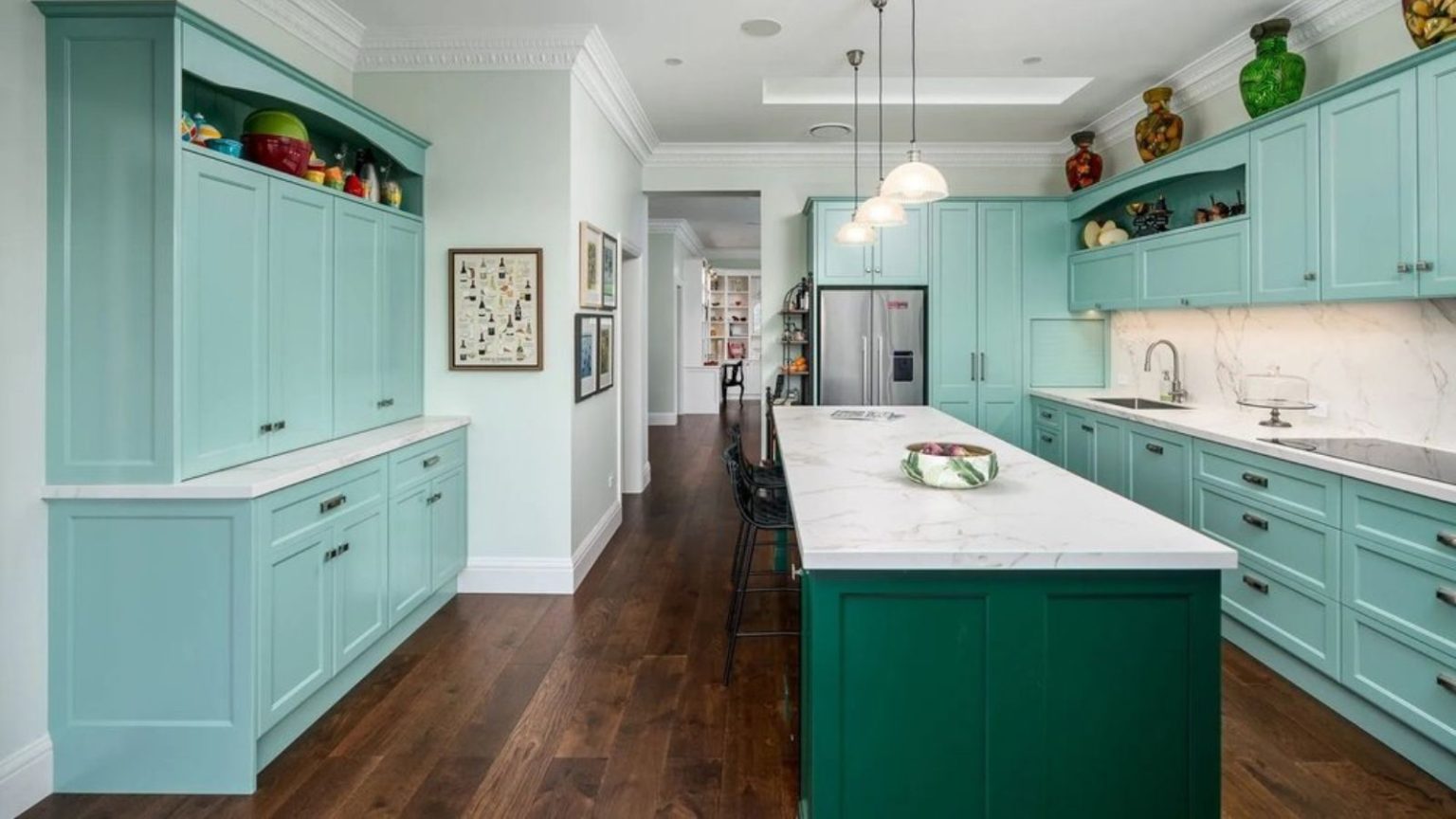Passez moins de temps à nettoyer avec un puissant aspirateur robot eufy qui le fait pour vous – profitez de cette rare remise de 415 $ tant qu’elle dure Teal and green kitchen with kitchen island, wooden floors, three hanging lights. Vases and ornaments are positioned on the shelves and above cabinets, and a hallway is pictured in the background.
