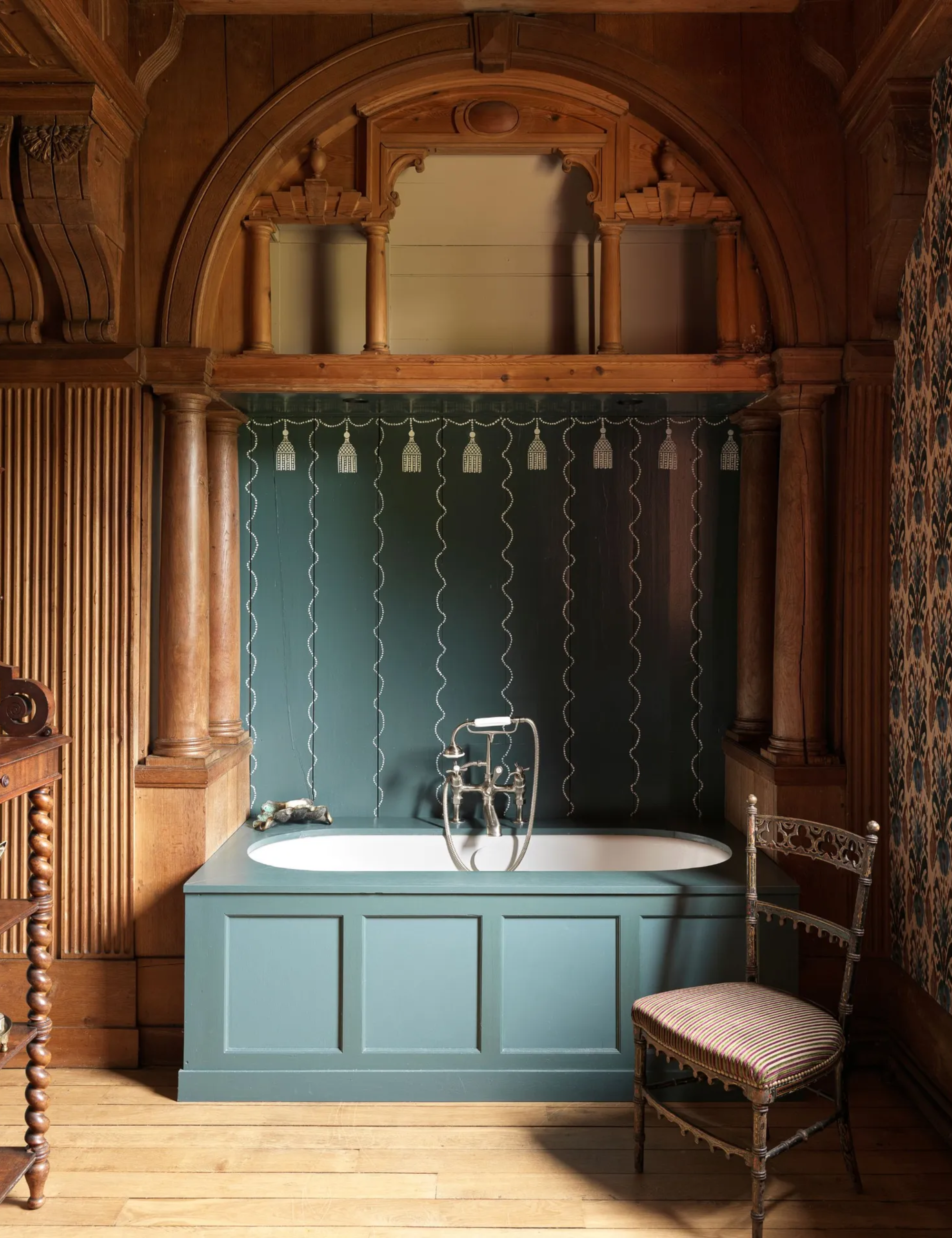 Une baignoire avec des panneaux extérieurs peints en bleu sarcelle avec des panneaux sur le mur derrière peints dans la même couleur bleu sarcelle audacieuse, avec des détails peints complexes. Il y a une chaise autonome au premier plan et un impressionnant plafond voûté.