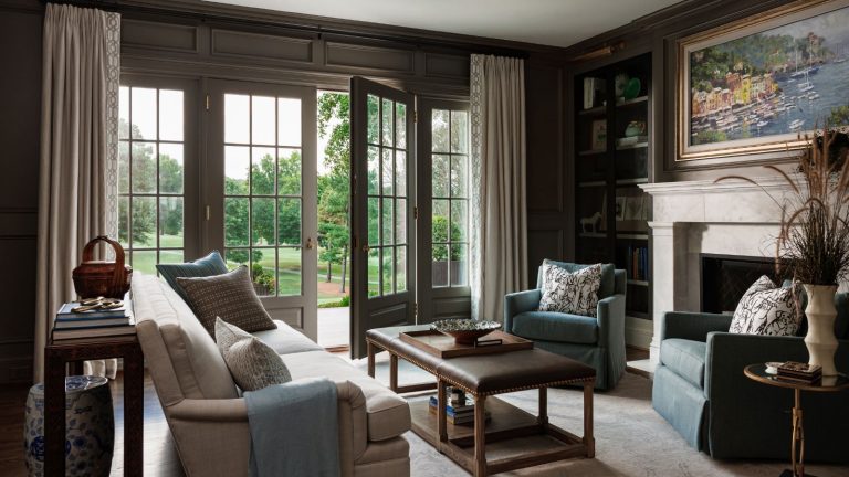 A large home office with dark gray-brown walls, French doors leading into a garden, and a cream sofa with an upholstered coffee table, and two blue armchairs in front of the fireplace.