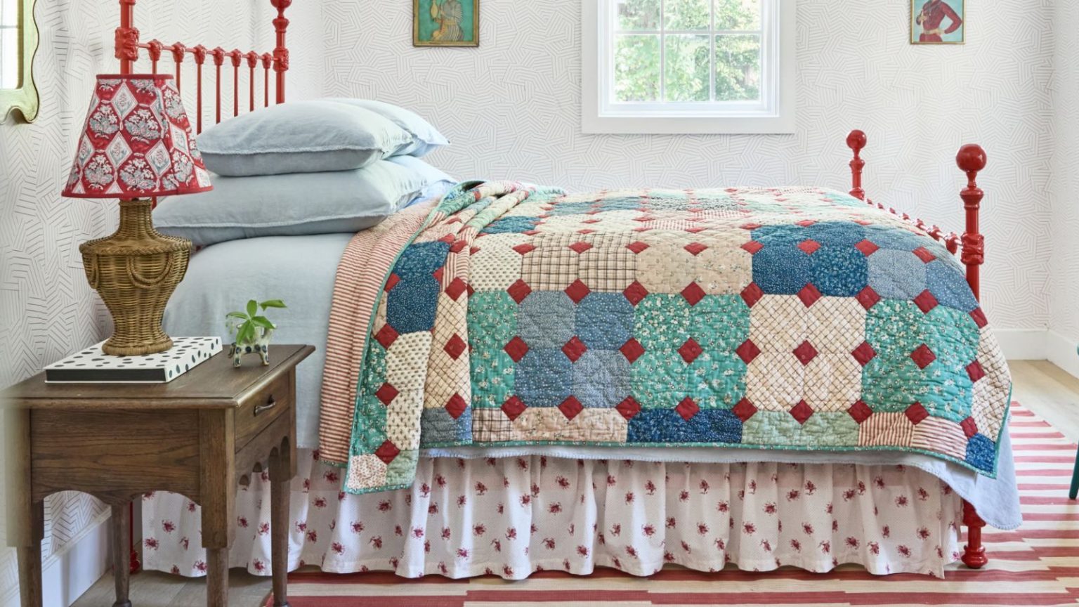 Ce commutateur de 2 minutes a juste déguisé mon rangement sous le lit et a instantanément donné à ma chambre un look plus fantaisiste – vous pouvez le récupérer pour seulement 50 $ a cottage style bedroom with a rug striped rug, red wrought iron bed, blue linens, a floral bed skirt and patchwork quilt with a wooden nightstand and rattan lamp
