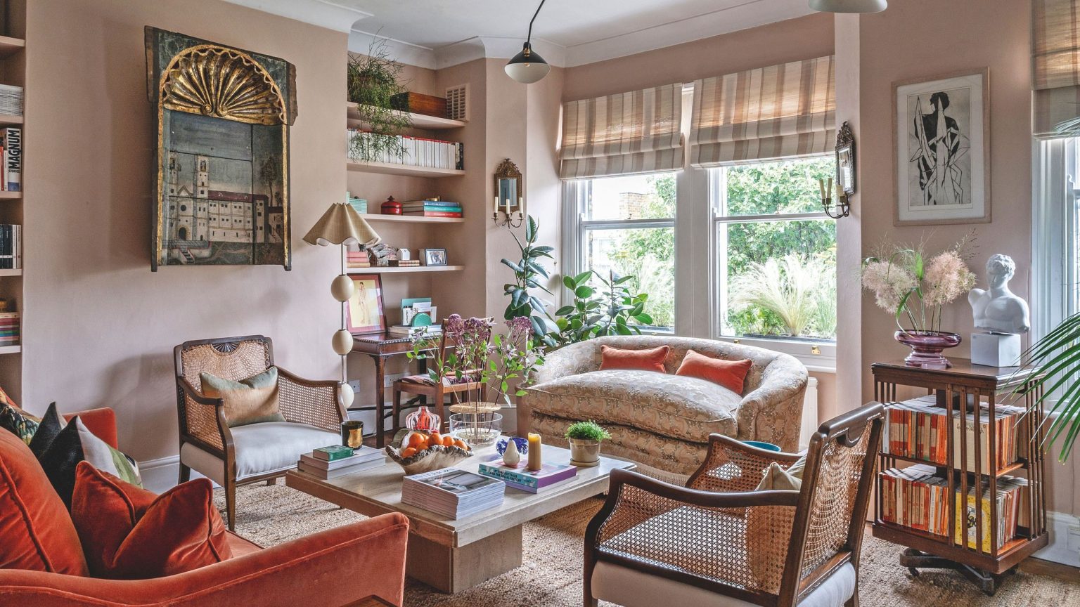 Dans ce petit appartement londonien, une cuisine IKEA minimaliste se mêle à des antiquités de caractère, des objets de famille et des œuvres d’art de collection – c’est une leçon de design haut-bas A warm, eclectic living room featuring dusty pink walls, a burnt orange velvet sofa, and woven cane armchairs arranged around a stone coffee table