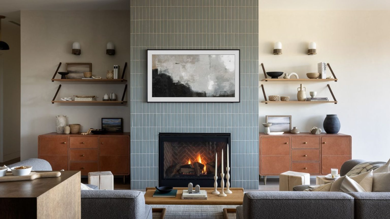 Les barres de son intégrées et personnalisées sont la prochaine grande tendance audio domestique – Voici pourquoi les experts sont divisés Living room with blue tiles and white walls, with floating shelves with various ornaments, a framed painting, a fireplace, two gray sofas, and a coffee table with books and candlesticks.