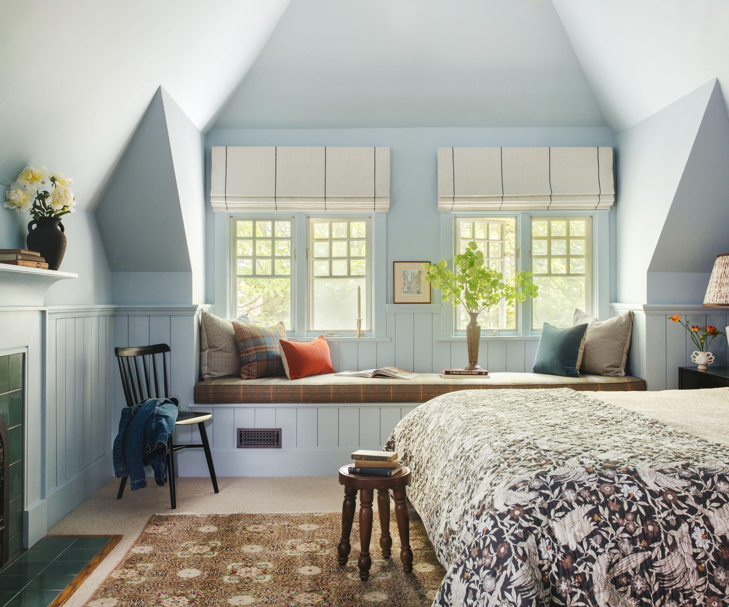 Chambre aux murs bleu pâle et cheminée carrelée vert foncé. Au centre se trouve un siège de fenêtre à panneaux avec des coussins et deux fenêtres. Le lit est doté d'un jeté fleuri et d'un tapis marron foncé en dessous.