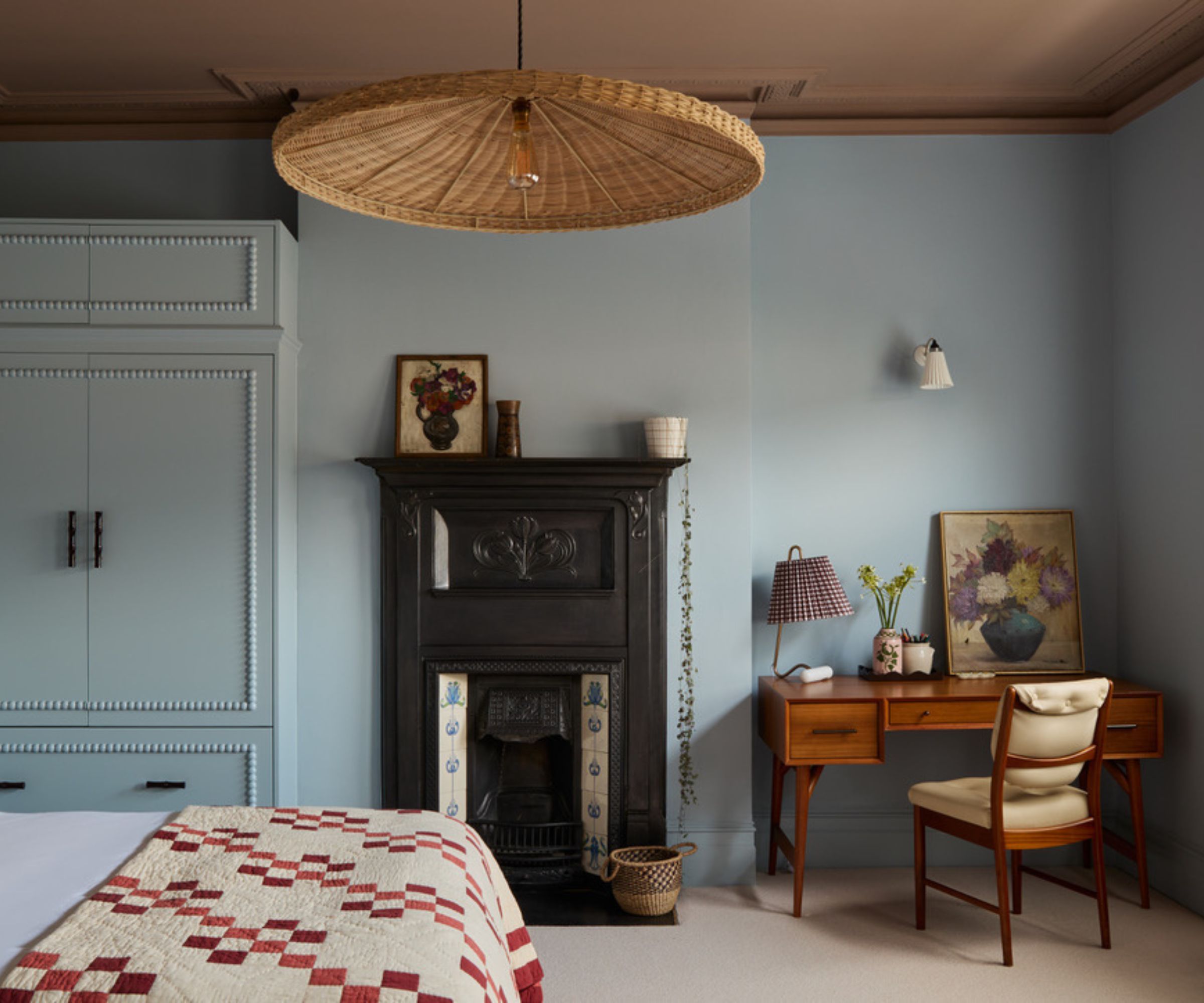 Chambre avec murs bleu pâle et plafond en plâtre rose, grande suspension en rotin, cheminée et bureau du milieu du siècle dans le coin avec des œuvres d'art et une lampe de table plissée. Un lit est doté d'une couette vintage rouge et crème.