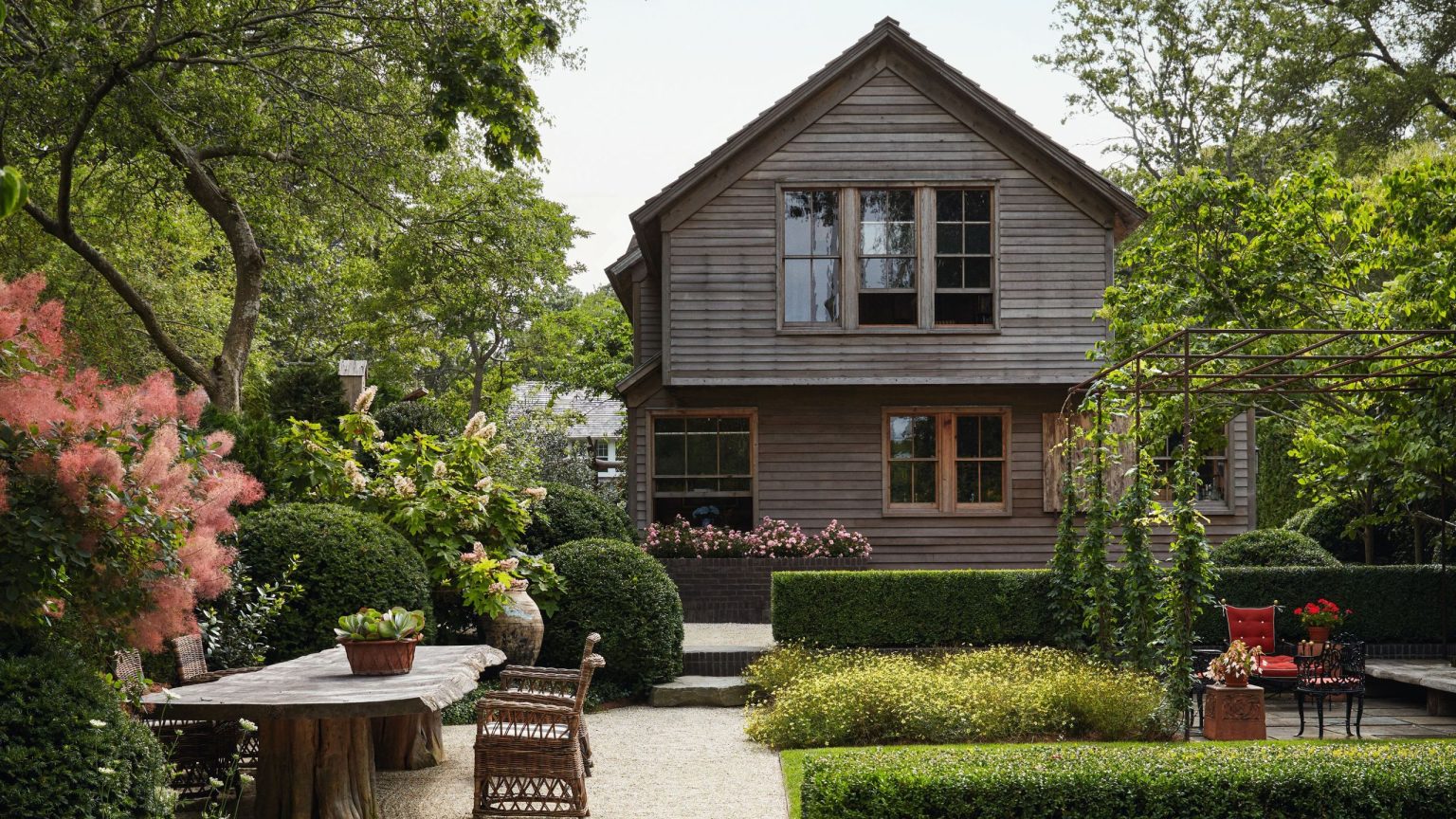 À l’intérieur d’une maison de Sag Harbor recouverte de cèdre qui équilibre les caractéristiques historiques avec un modernisme chaleureux A lush, tiered backyard garden with manicured hedges, stone pathways, and a large outdoor wooden dining table. In the background stands a weathered two-story grey shingle house surrounded by mature trees.