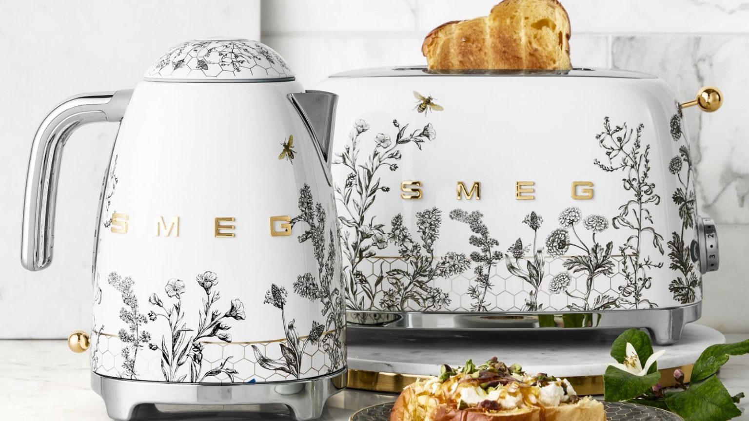 Appareils de décoration en nid d’abeille ? La tendance fantaisiste de la cuisine 2026 que vous n’avez pas vue venir A SMEG kettle and toaster with grey flower and bee motifs on white bases, with stainless steel accents.